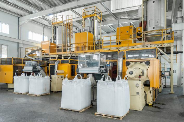 Linha de produção industrial com máquinas automatizadas usadas no processamento de plásticos. Quatro grandes sacos brancos (big bags) estão cheios de matéria-prima, possivelmente grânulos de polipropileno. O ambiente é limpo, moderno e bem iluminado.








