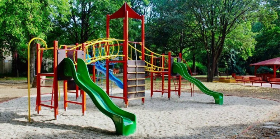 Uma estrutura de parque infantil no centro de um espaço ao ar livre, com escorregadores verdes, plataformas e barras de apoio. O solo ao redor é coberto por areia, e há árvores verdes ao fundo, proporcionando sombra e um ambiente natural. Ao fundo, também é possível ver bancos de madeira e uma área de descanso com toldo.

